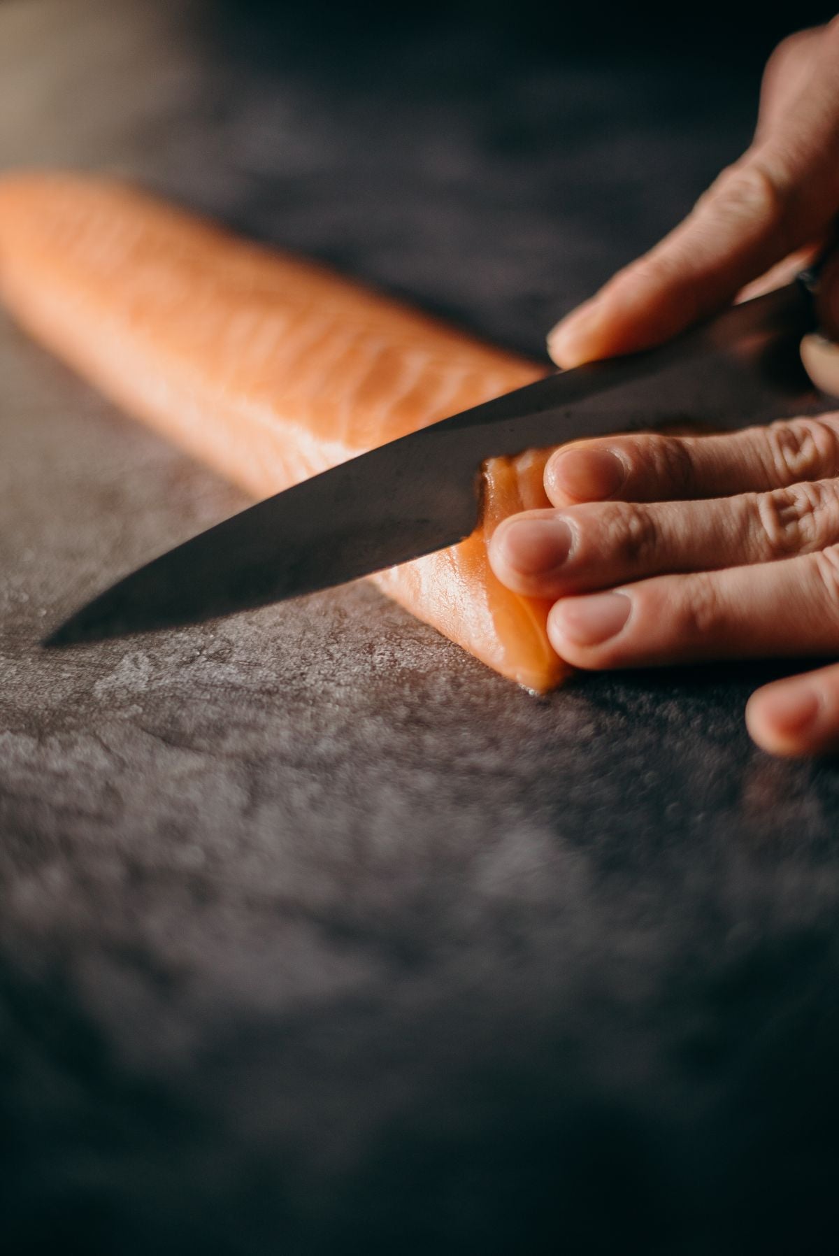 Raw salmon fillet being sliced on a dark stone surface